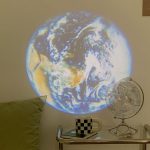 Projeção do planeta Terra na parede com detalhes visíveis de continentes e oceanos, iluminada por luz azul e branca, com um globo terrestre real ao lado.