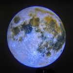Fotografia da lua mostrando crateras, mares e relevo lunar com detalhes visíveis, iluminada por luz solar, contra fundo escuro do espaço.