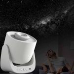 Projetor de estrelas branco a projetar o céu noturno na parede, com uma pessoa a observar e uma criança deitada na cama, ambiente escuro.