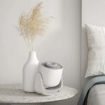 Humidificador de ar branco com design elegante, colocado numa mesa de pedra junto a uma planta seca e uma cama com almofadas.