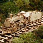 Locomotiva de madeira detalhada com rodas, chaminé e carruagem, sobre trilhos de madeira, rodeada de vegetação verde.