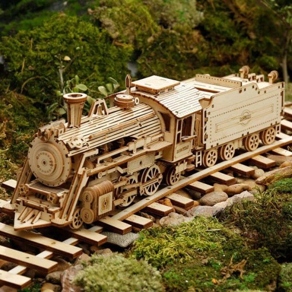 Locomotiva de madeira detalhada com rodas, chaminé e carruagem, sobre trilhos de madeira, rodeada de vegetação verde.
