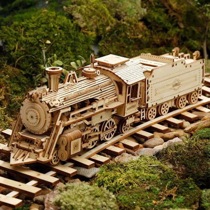 Locomotiva de madeira detalhada com rodas, chaminé e carruagem, sobre trilhos de madeira, rodeada de vegetação verde.