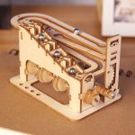 Puzzle mecânico de madeira com várias peças e bolas metálicas, incluindo uma rampa e engrenagens, sobre uma superfície de madeira clara.