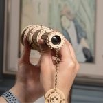 Visão detalhada de um telescópio 3D de madeira com partes ajustáveis e lente, segurado por uma mão, com fundo de parede com pintura de flamingo.