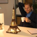 Modelo de torre Eiffel em miniatura com iluminação LED, visto de frente, com detalhes arquitetónicos visíveis, colocado numa mesa de escritório com um homem sorridente ao lado.
