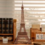 Miniatura da Torre Eiffel luminosa com detalhes intricados, colocada numa mesa de estudo com livros e calendário de agosto ao fundo.