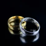 Anéis de compromisso dourado e prateado com detalhes gravados, colocados sobre uma superfície escura, refletindo a luz.