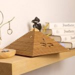 Quebra-cabeças em forma de pirâmide de madeira com várias peças encaixadas, sobre uma mesa de madeira clara, com livros e decoração ao fundo.