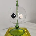 Radiómetro de Crookes com esfera de vidro transparente, lâmina de papel e lâmina de metal, montado em suporte de vidro verde, sobre base amarela.