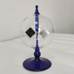 Radiómetro de Crookes com esfera de vidro transparente, hélice azul e dois espelhos, um preto e um prateado, montados em suporte azul.