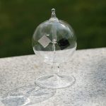 Radiómetro de Crookes em vidro transparente com lâmpada de plasma visível no interior, colocado numa superfície de pedra ao ar livre.