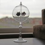 Radiómetro solar com esfera de vidro transparente, hélices pretas e suporte de vidro, colocado numa mesa junto a uma janela com vista para o exterior.