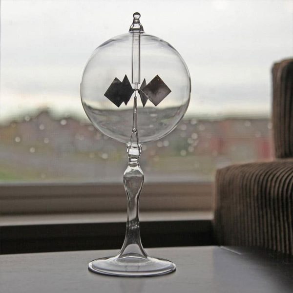 Radiómetro solar com esfera de vidro transparente, hélices pretas e suporte de vidro, colocado numa mesa junto a uma janela com vista para o exterior.