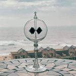 Radiómetro solar transparente com duas pás pretas, colocado numa superfície de mosaico, com vista para o mar e casas ao fundo.