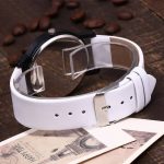 Relógio com pulseira branca, visor transparente e ponteiros invertidos, colocado sobre uma mesa de madeira com café ao fundo.