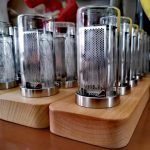 Conjunto de relógios de tubo luminoso com estrutura de vidro e base de madeira, exibidos em fila numa loja de gadgets científicos.