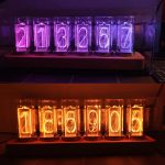 Relógio de tubo luminoso com números de cores diferentes, exibindo horas e minutos em dois conjuntos distintos, com iluminação vibrante em tons de roxo, azul e laranja.
