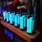 Conjunto de relógios de tubo luminoso com números azuis, exibidos numa base de madeira, com fundo decorativo de brinquedos de ciência.