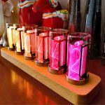Conjunto de relógios de tubo luminoso com diferentes cores de luz, exibidos numa base de madeira, com detalhes em neon rosa, azul, amarelo, laranja, vermelho e verde.