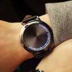 Relógio digital com mostrador circular e luzes LED azuis ao redor, usado no pulso de uma pessoa com roupa cinza e pele clara.