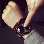Pessoa usando um relógio inteligente com mostrador digital, apontando para a tela com o dedo, usando pulseira de beads vermelhos e roupa cinza.
