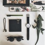 Relógio de pulso com bracelete preto, óculos escuros redondos, bloco de notas preto com clipe, caneta verde, colar de corrente com cadeado, lagarto de brinquedo de plástico, e caixas com ilustrações.