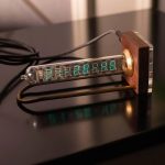 Relógio Nixie com tubo de vidro visível, exibindo hora digital em números verdes, com base de madeira e componentes elétricos visíveis.