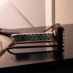 Relógio Nixie com visor de tubo de vidro exibindo hora, com iluminação fluorescente azul e suporte de madeira escura.