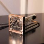 Módulo de relógio Nixie com componentes eletrónicos visíveis, caixa de acrílico transparente e circuito impresso exposto, com detalhes de componentes internos e estrutura metálica.