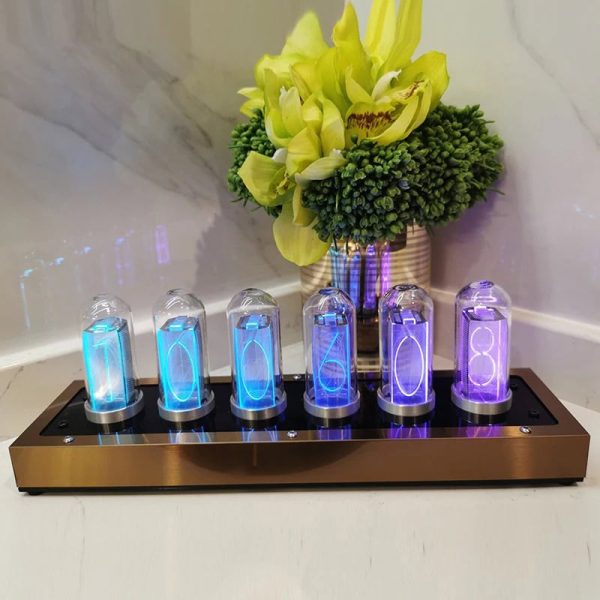 Relógio Nixie com tubos de vidro iluminados em azul e roxo, colocado numa base de metal, com vaso de flores amarelas e verdes ao fundo.