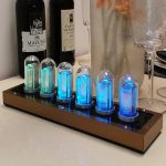 Relógio Nixie com tubos de vidro iluminados em cores diferentes, colocado numa base de metal, com garrafas de vinho e copos ao fundo.