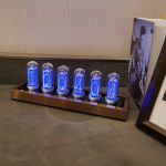 Relógio Nixie com tubos de vidro transparentes, exibindo números azuis brilhantes, montado numa base de metal com iluminação LED azul.