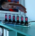 Relógio Nixie com tubos de vidro exibindo números vermelhos, com iluminação RGB ajustável, colocado numa base transparente sobre uma mesa, com um computador portátil ao fundo.