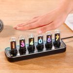Relógio Nixie com tubos de vidro numerados de 1 a 7, exibindo números coloridos, colocado numa base preta sobre uma mesa de madeira.
