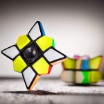 Cubo de Rubik com peças coloridas, destaque para uma face com cores amarelo, branco e preto, com fundo desfocado de outros brinquedos de ciência.