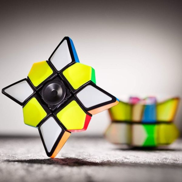 Cubo de Rubik com peças coloridas, destaque para uma face com cores amarelo, branco e preto, com fundo desfocado de outros brinquedos de ciência.