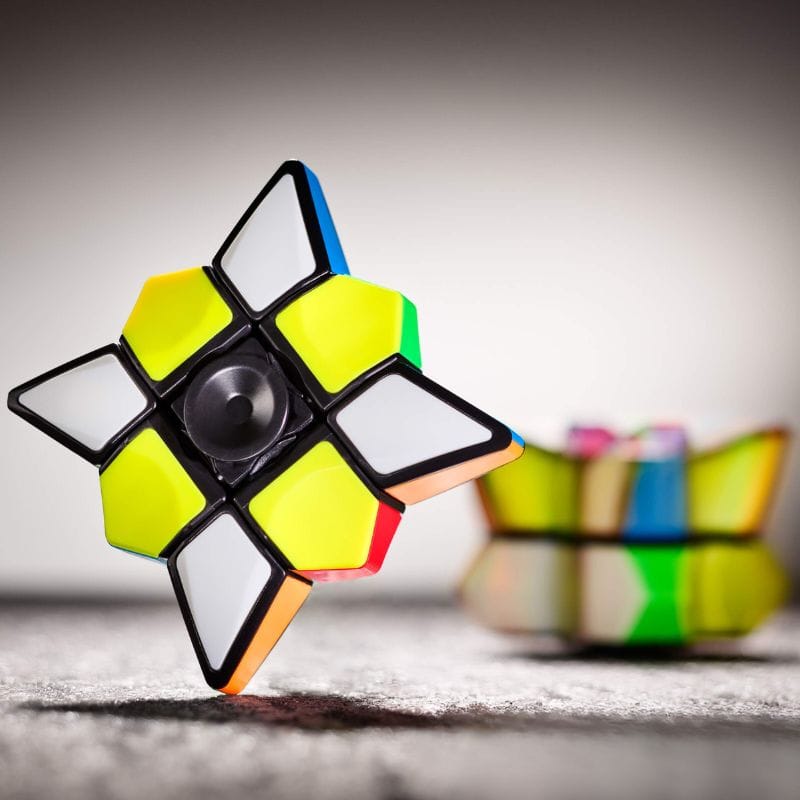 Cubo de Rubik com peças coloridas, destaque para uma face com cores amarelo, branco e preto, com fundo desfocado de outros brinquedos de ciência.