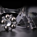 Spinner de mão com várias esferas de metal brilhantes e estrutura de suporte de metal cinzento, colocado sobre uma superfície escura com fundo desfocado.