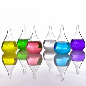 Seis frascos de vidro com líquidos de cores diferentes: amarelo, verde, rosa, transparente, azul e roxo, refletidos numa superfície brilhante.