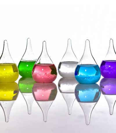 Seis frascos de vidro com líquidos de cores diferentes: amarelo, verde, rosa, transparente, azul e roxo, refletidos numa superfície brilhante.