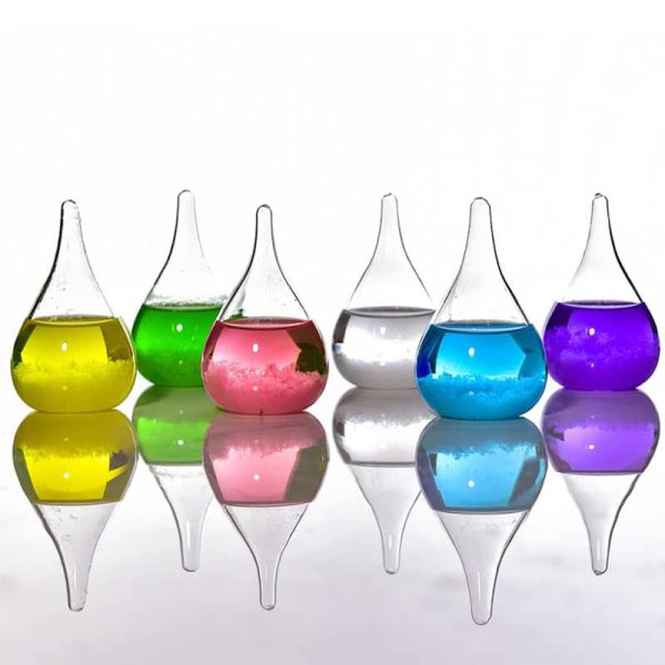 Seis frascos de vidro com líquidos de cores diferentes: amarelo, verde, rosa, transparente, azul e roxo, refletidos numa superfície brilhante.