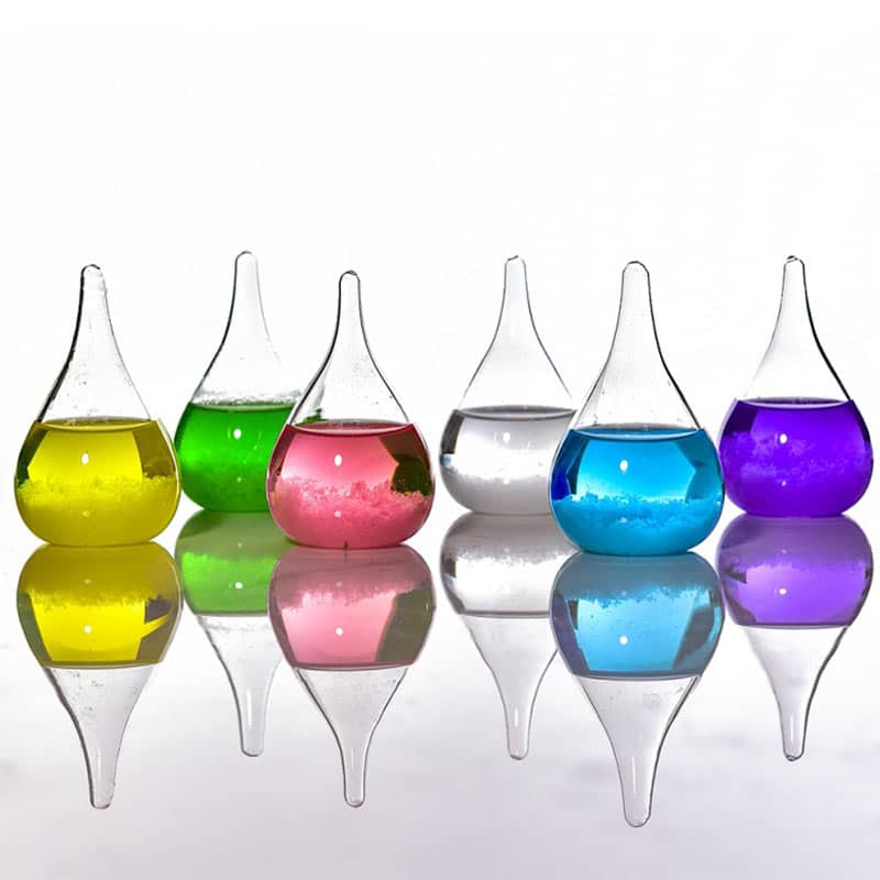 Seis frascos de vidro com líquidos de cores diferentes: amarelo, verde, rosa, transparente, azul e roxo, refletidos numa superfície brilhante.