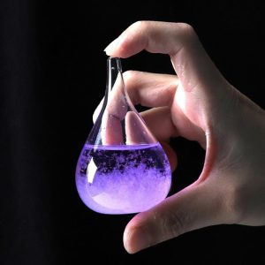 Frasco de vidro em forma de lágrima com líquido roxo e efeito de bolhas, segurado por uma mão contra fundo escuro.