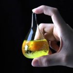 Frasco de vidro em forma de lágrima com líquido amarelo fluorescente e bolhas, segurado por uma mão contra fundo escuro.