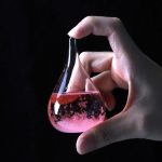 Frasco de vidro em forma de lágrima com líquido rosa e bolhas, segurado por uma mão, fundo escuro.