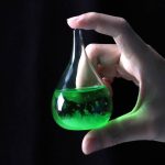 Frasco de vidro em forma de lágrima com líquido verde fluorescente, segurado por uma mão, fundo preto.