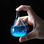 Frasco de vidro com líquido azul brilhante, sendo segurado por uma mão, fundo preto.