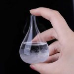 Frasco de vidro em forma de lágrima com líquido transparente e vapor, sendo segurado por uma mão, fundo preto.