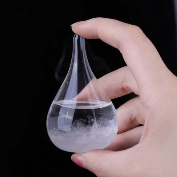 Frasco de vidro em forma de lágrima com líquido transparente e vapor, sendo segurado por uma mão, fundo preto.